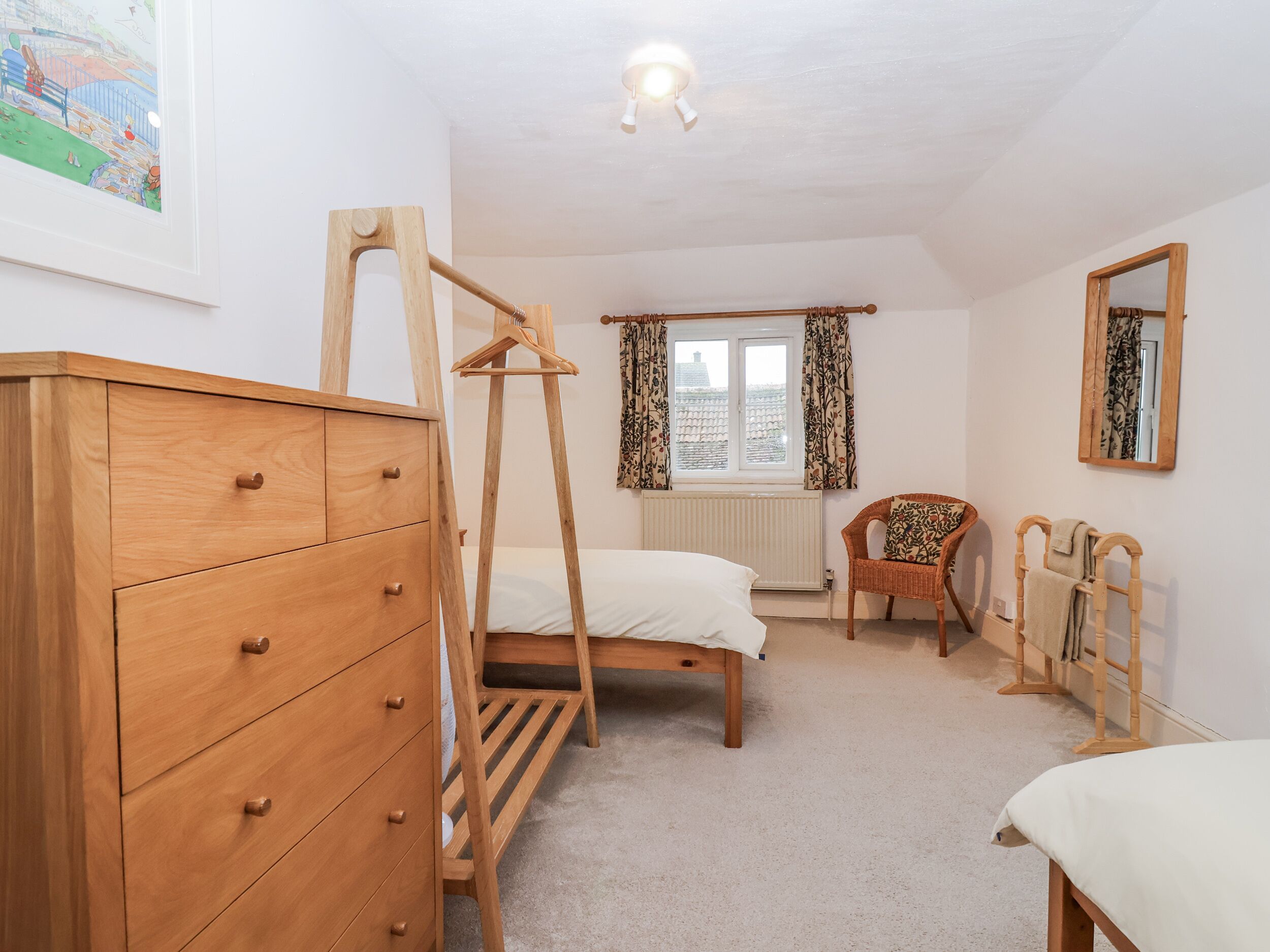 Un dormitorio con un aparador de madera, una cama con estructura de madera, una silla, un tendedero de madera y una ventana con cortinas.
