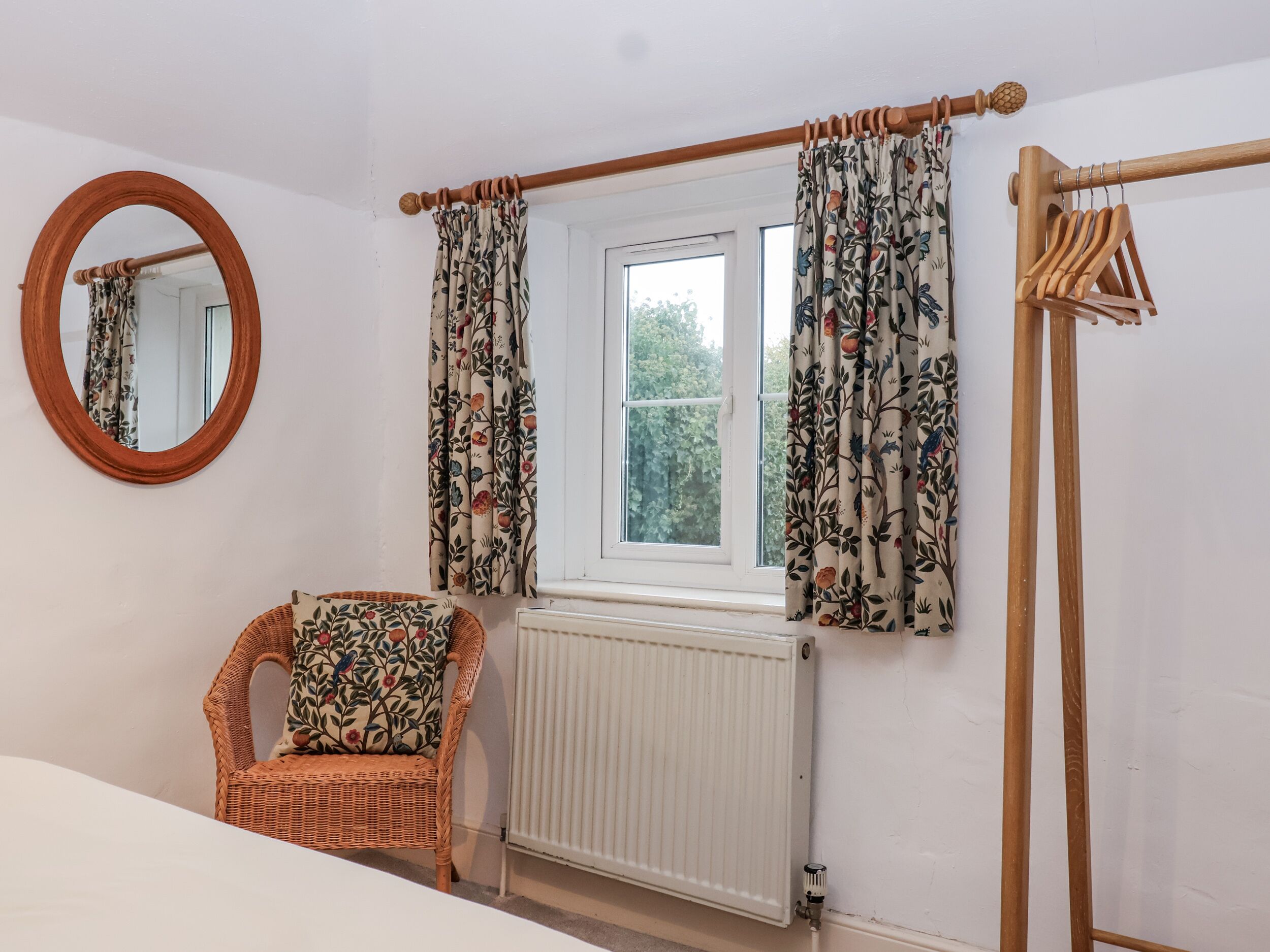 Een slaapkamer met een rieten stoel, een ronde spiegel, een raam met bloemen gordijnen en een radiator.