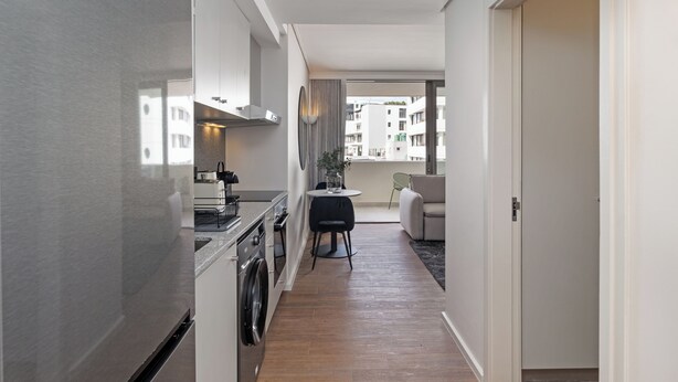 A narrow hallway with a kitchen on one side, a dining area with a table and chairs, and a view of buildings outside. - Dolce Vita | Stylish Sea Point Apartment (Cape Town)
