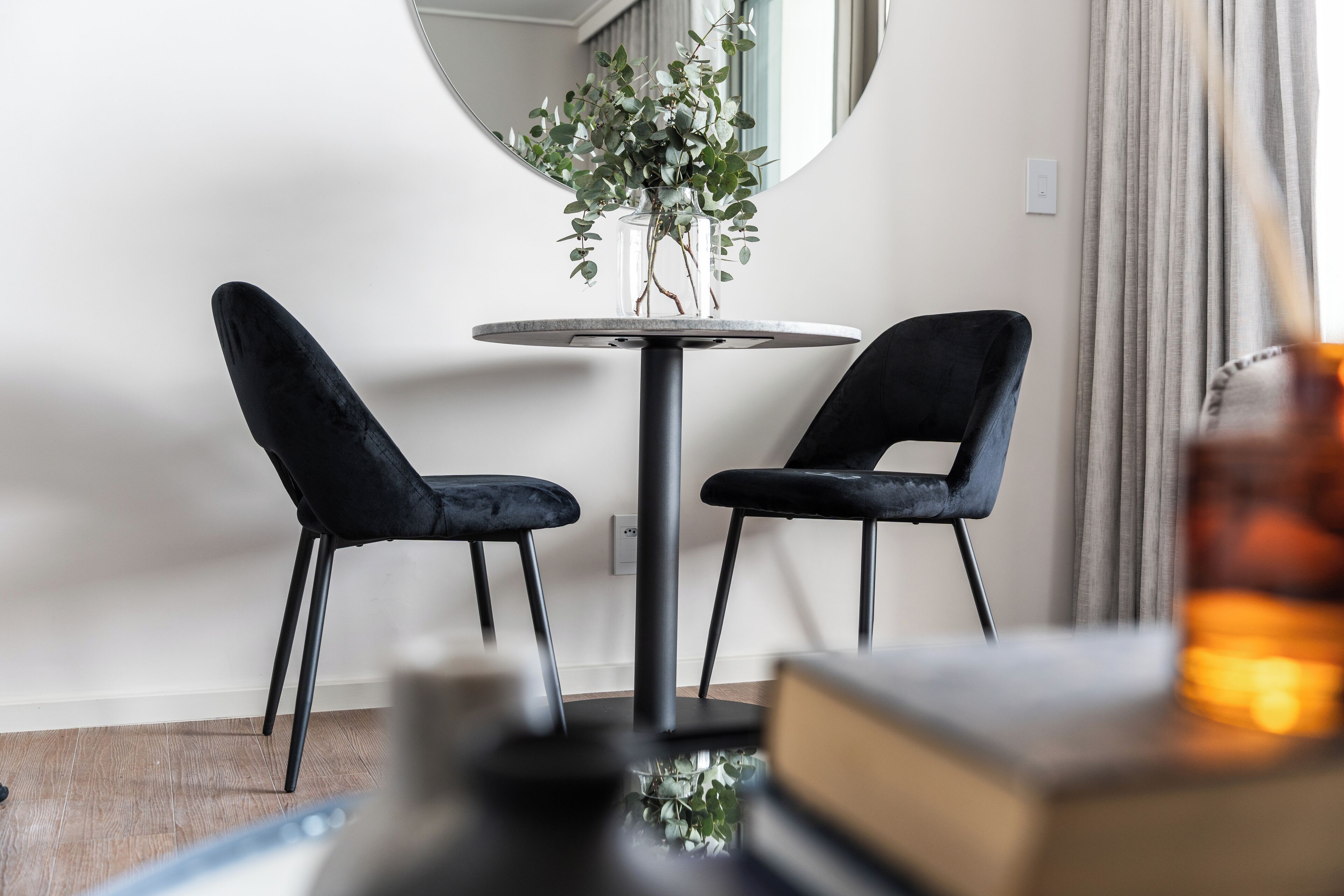 Een moderne kamer met twee zwarte stoelen rond een ronde tafel, een spiegel aan de muur en een plant in een glazen vaas.