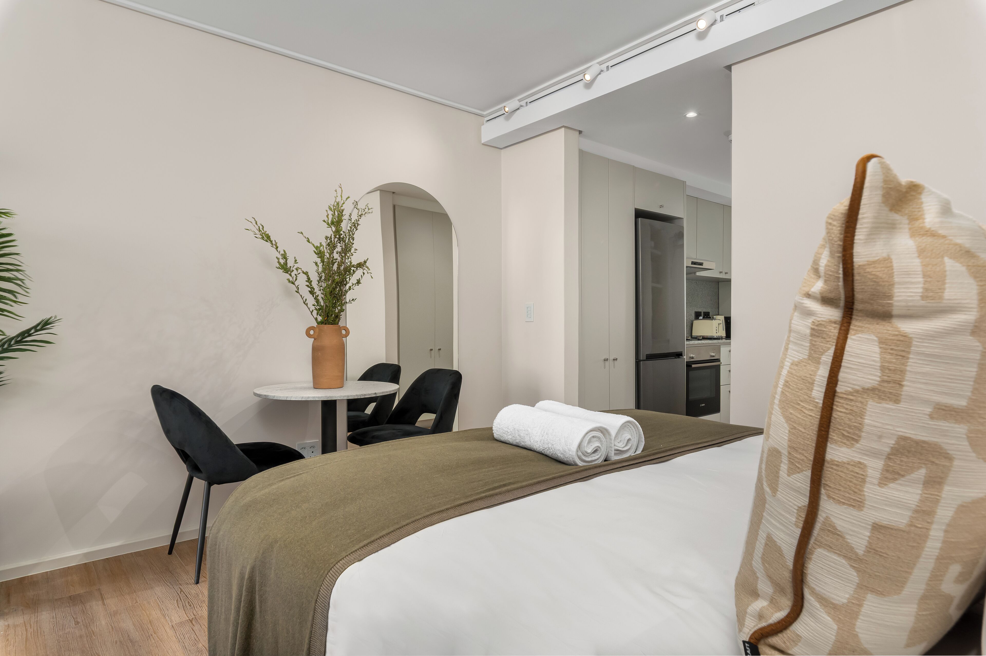 A hotel room with a bed, a desk, a chair, a small table, and a potted plant.