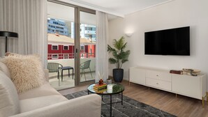 Una sala moderna con un sofá, una mesa de centro con libros, una planta en maceta, un televisor y un ventanal corredero que da a un balcón exterior.