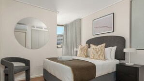 Habitación de hotel moderna con una cama grande, un espejo redondo, una silla, una mesita de noche con lámpara y vistas a la ciudad a través de la ventana.