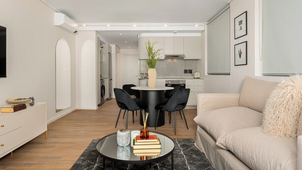 Una sala moderna con una mesa de centro redonda de vidrio, un sofá color beige y un comedor con sillas negras y una mesa blanca.
