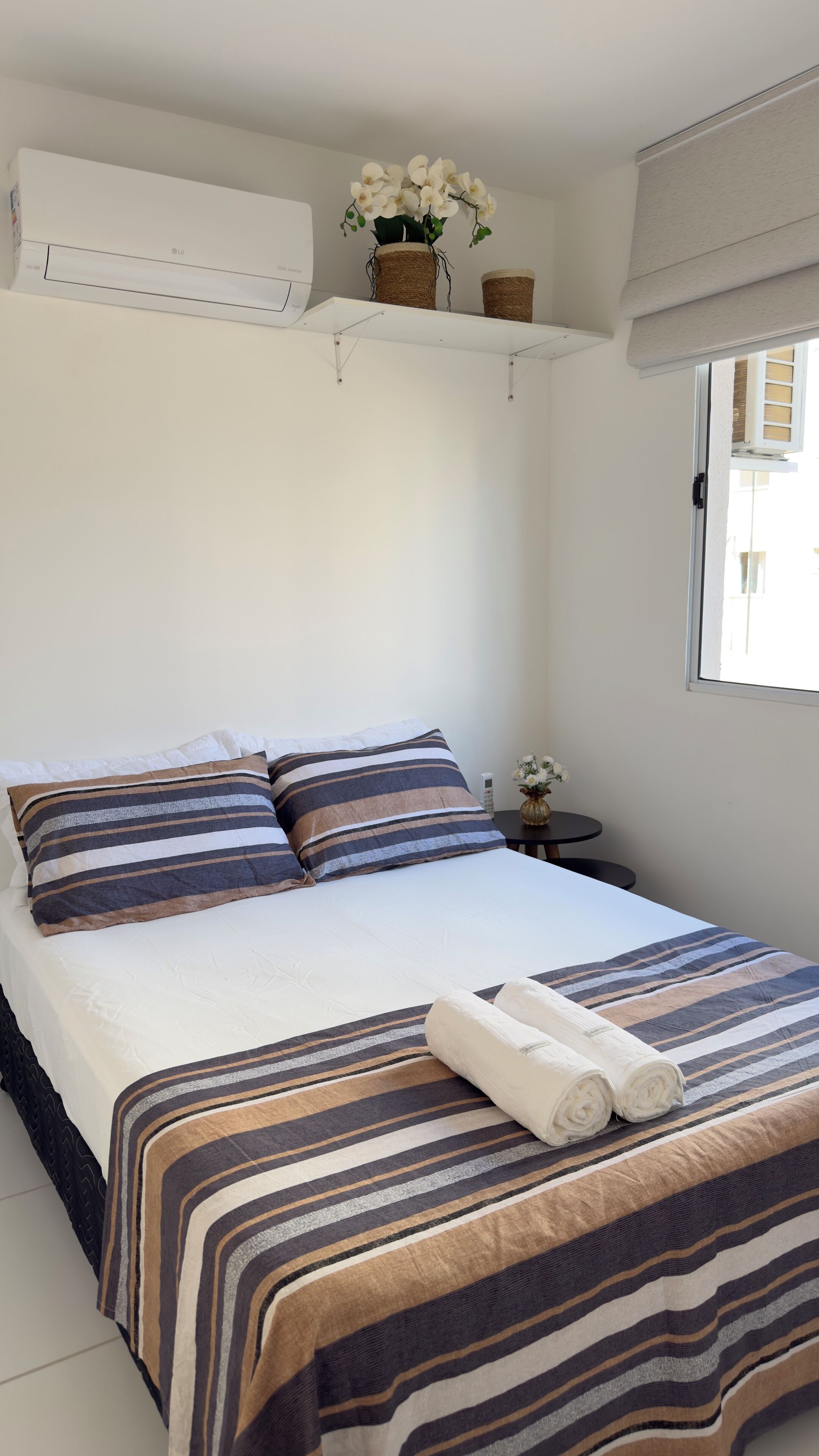 A neatly made bed with striped bedding, a rolled towel, and a small table with a vase of flowers.