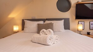 Een netjes opgemaakt bed met opgerolde handdoeken, een ronde wandklok en een flatscreen televisie aan de muur.