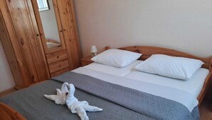 Een netjes opgemaakt bed met witte kussens en een grijze deken, een houten garderobe en een nachtkastje met een lamp.