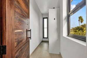 A hallway with a wooden door, a white wall, a window with a view of palm trees, and a door leading to another room.