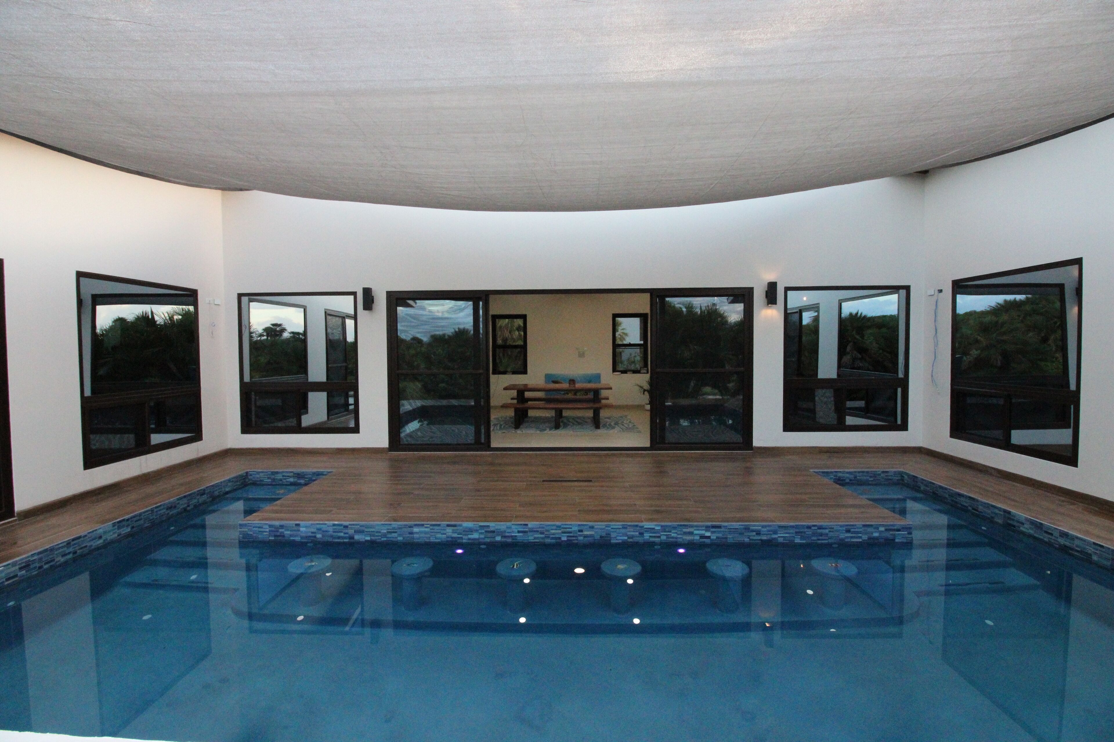 Une piscine intérieure moderne avec une terrasse en bois, de grandes portes vitrées et un banc à l’arrière-plan.