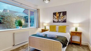 Een slaapkamer met een bed, twee nachtkastjes, een radiator, een raam met uitzicht op een huis en groen, en een kunstwerk aan de muur.