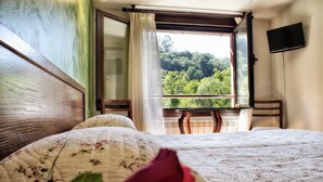 Un dormitorio con un ventanal grande, una cama con ropa de cama floral y vistas a una zona boscosa.