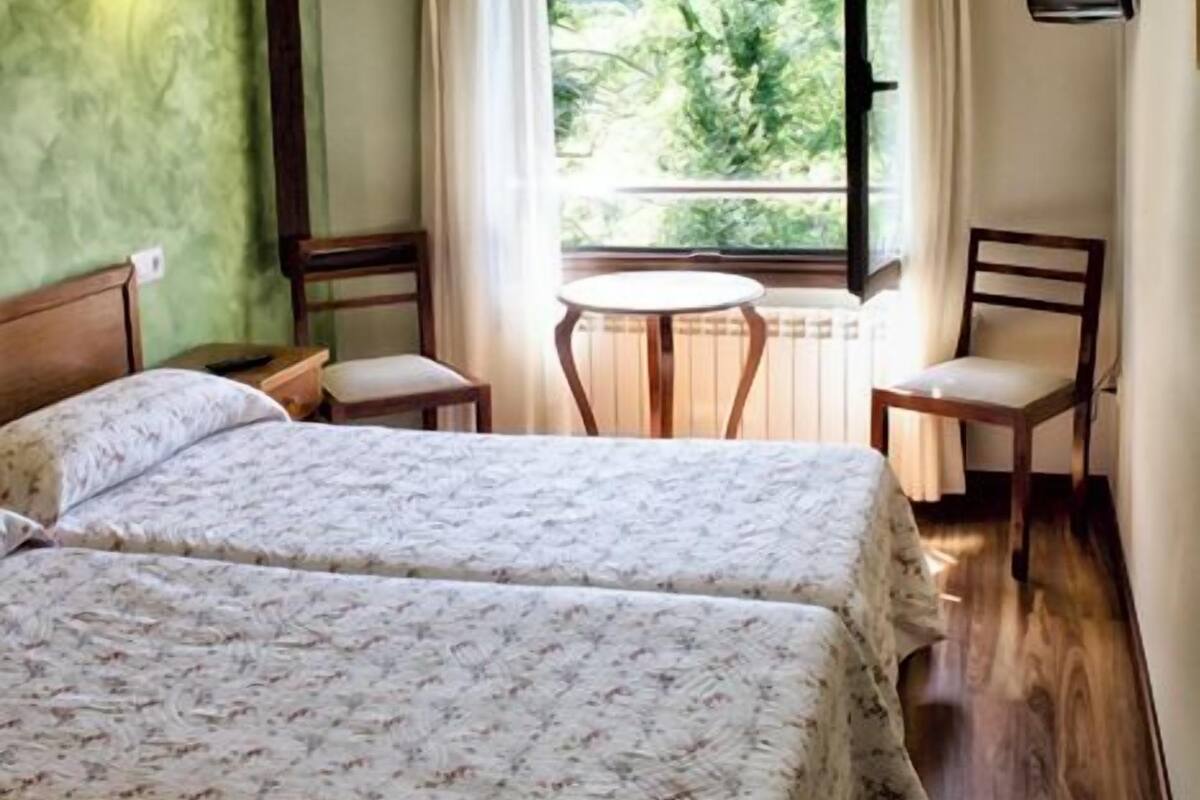 A bedroom with two beds, a window with curtains, a small table, and a chair.