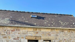 Ein Steinhaus mit einer dunklen Tür, zwei kleinen Fenstern und einer Schotterauffahrt.