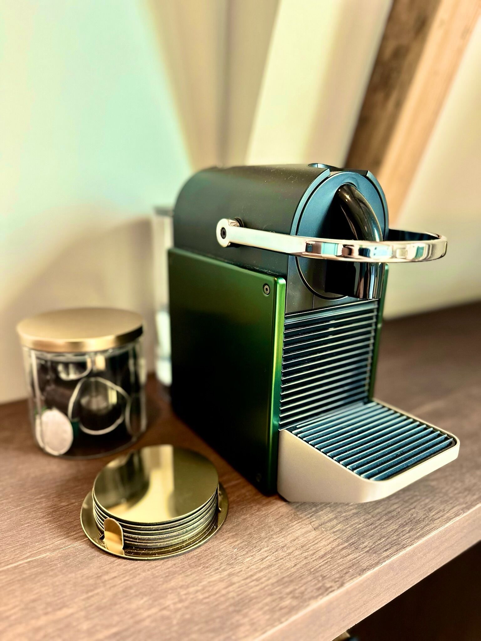 Eine kleine, grün-weiße Espressomaschine mit einem Metallhebel auf der Oberseite, auf einer Holzfläche neben einem Glasbehälter mit Deckel und einer Stapel Metallteller.