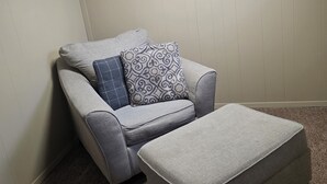 Habitación de esquina con sofá y reposapiés grises, un cojín estampado y un techo inclinado con tragaluces.