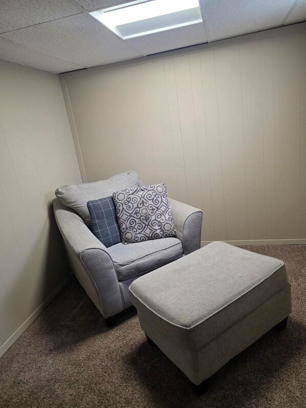 A corner room with a grey sofa and ottoman, a patterned pillow, and a slanted ceiling with skylight.