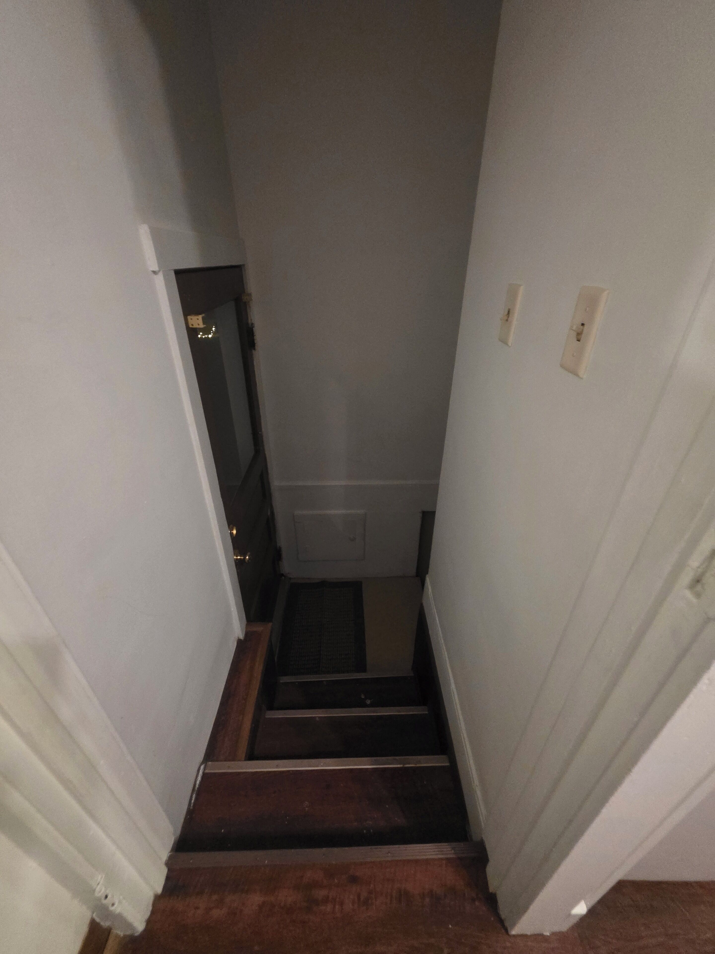 Eine schmale Treppe mit Holzboden und weißen Wänden führt zu einer Zwischenebene mit einem Lüftungsgitter.