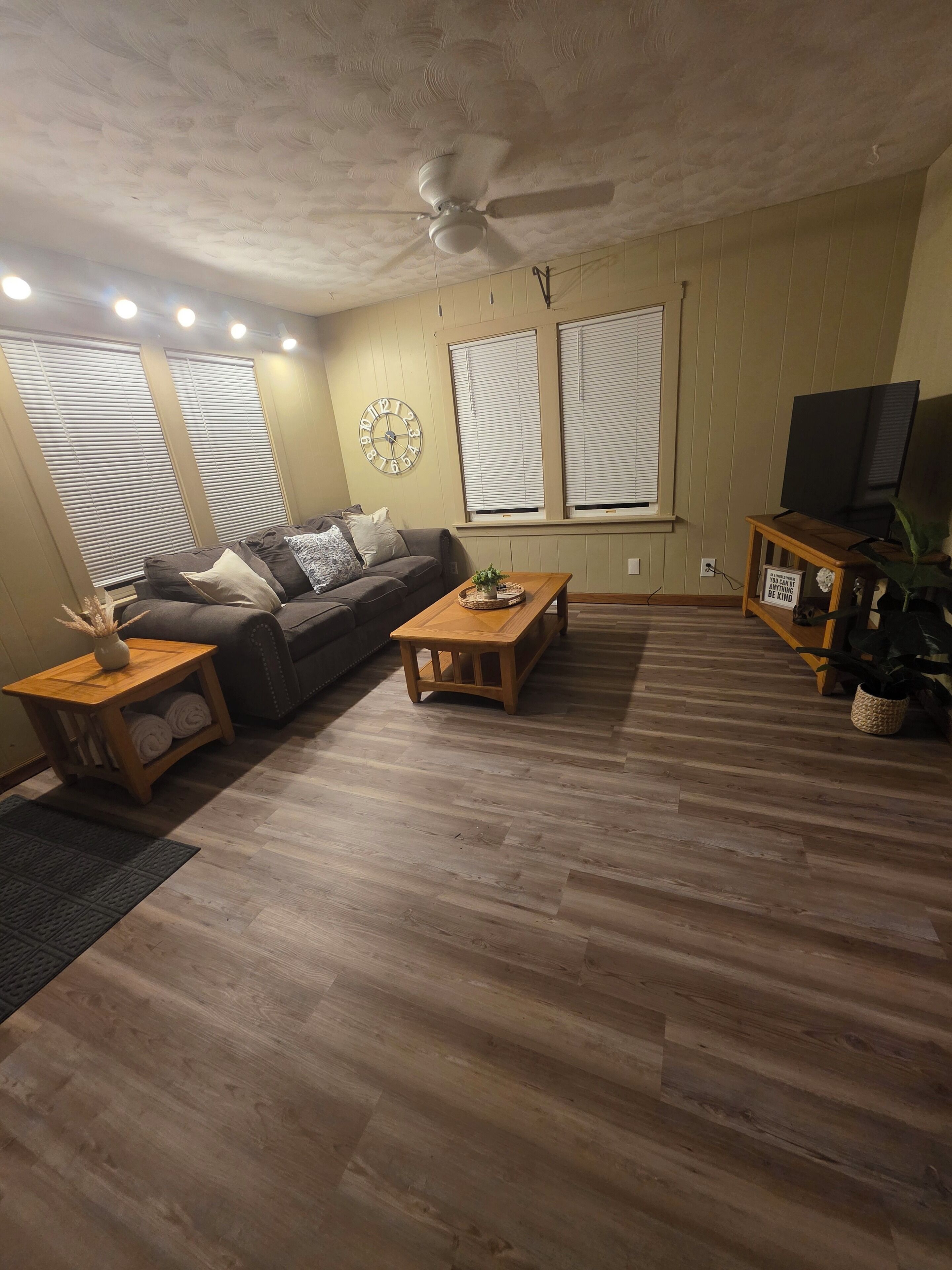 A living room with a wooden floor, a ceiling fan, a sofa, a coffee table, a side table with towels, a clock, and a TV on a stand.