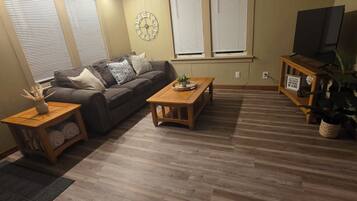 A living room with a wooden floor, a ceiling fan, a sofa, a coffee table, a side table with towels, a clock, and a TV on a stand.