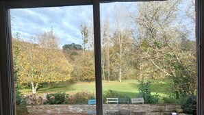 A view through a glass door of a patio area with a wooden deck, a round table, a blue chair, and a barbecue grill. - CLOS DU PIN - 6 pers ( Gite N°2 du domaine du Pin) (LANGUIDIC)