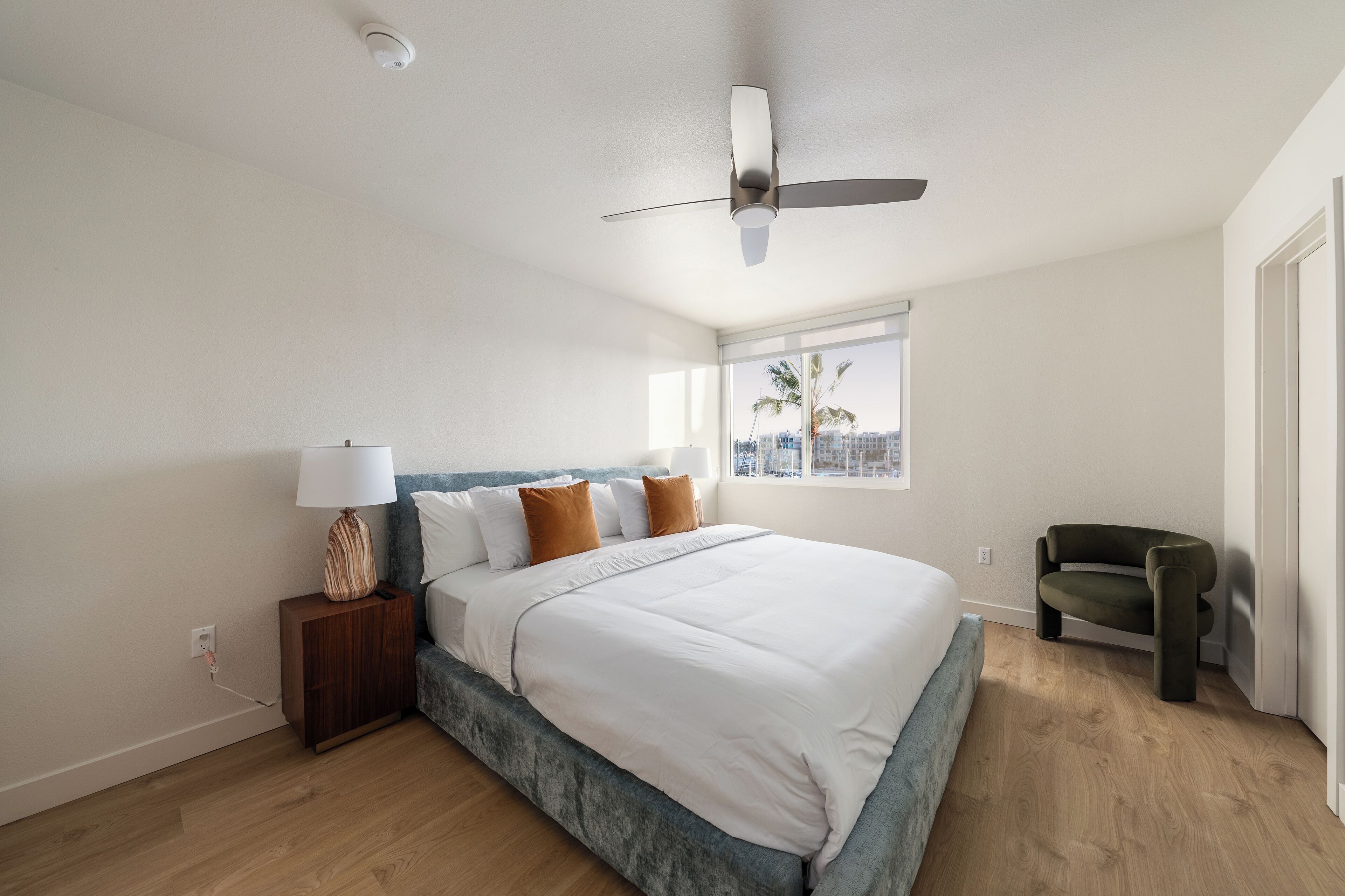 A bedroom with a bed, a nightstand, a lamp, a ceiling fan, a window with a view, and a chair.