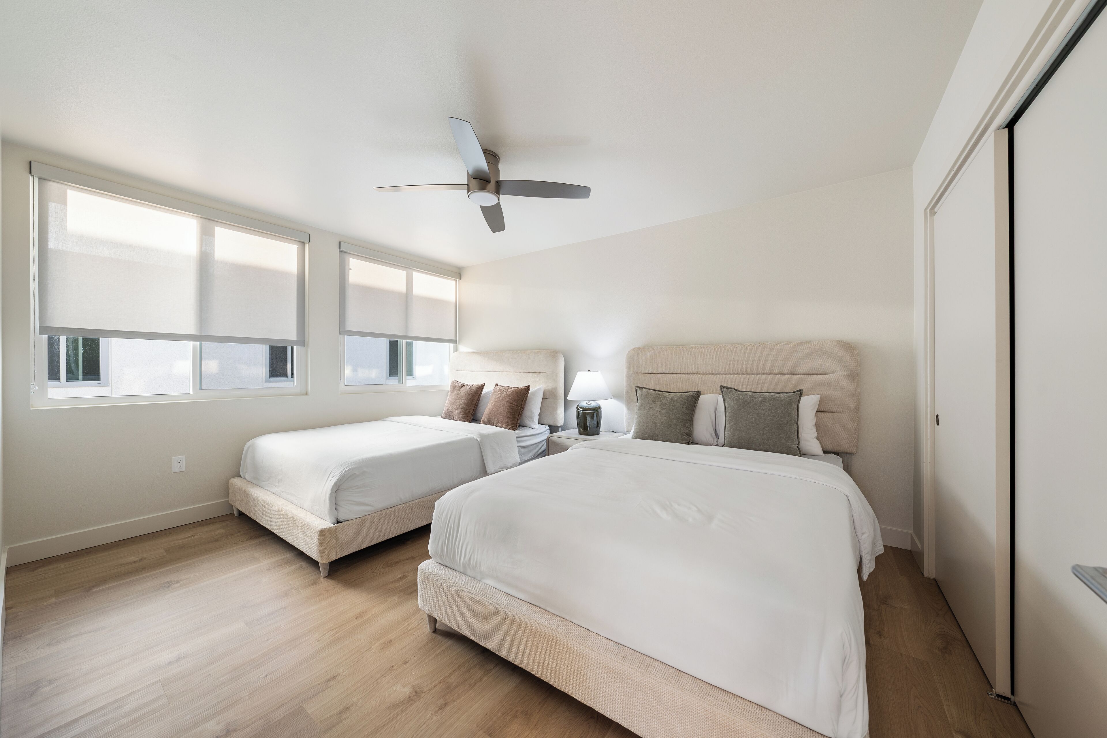 A bedroom with two beds, a ceiling fan, a bedside table with a lamp, and large windows.