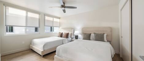 A bedroom with two beds, a ceiling fan, a bedside table with a lamp, and large windows.