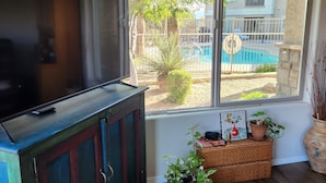 Une pièce avec un téléviseur posé sur un meuble, une fenêtre donnant sur une piscine et une plante en pot sur le rebord de la fenêtre.