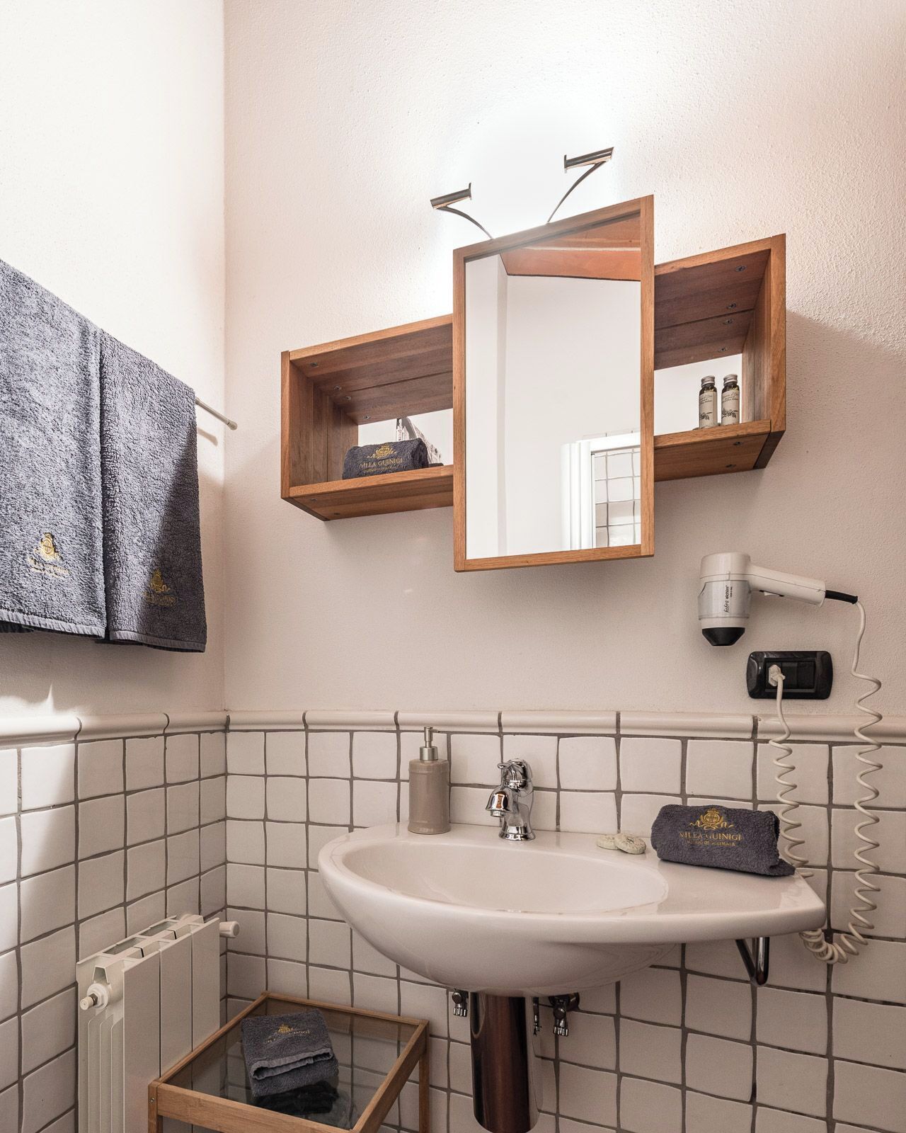 Une salle de bain avec un lavabo blanc, un miroir, un porte-serviettes, un sèche-cheveux et une petite étagère en bois où sont rangés les articles de toilette.