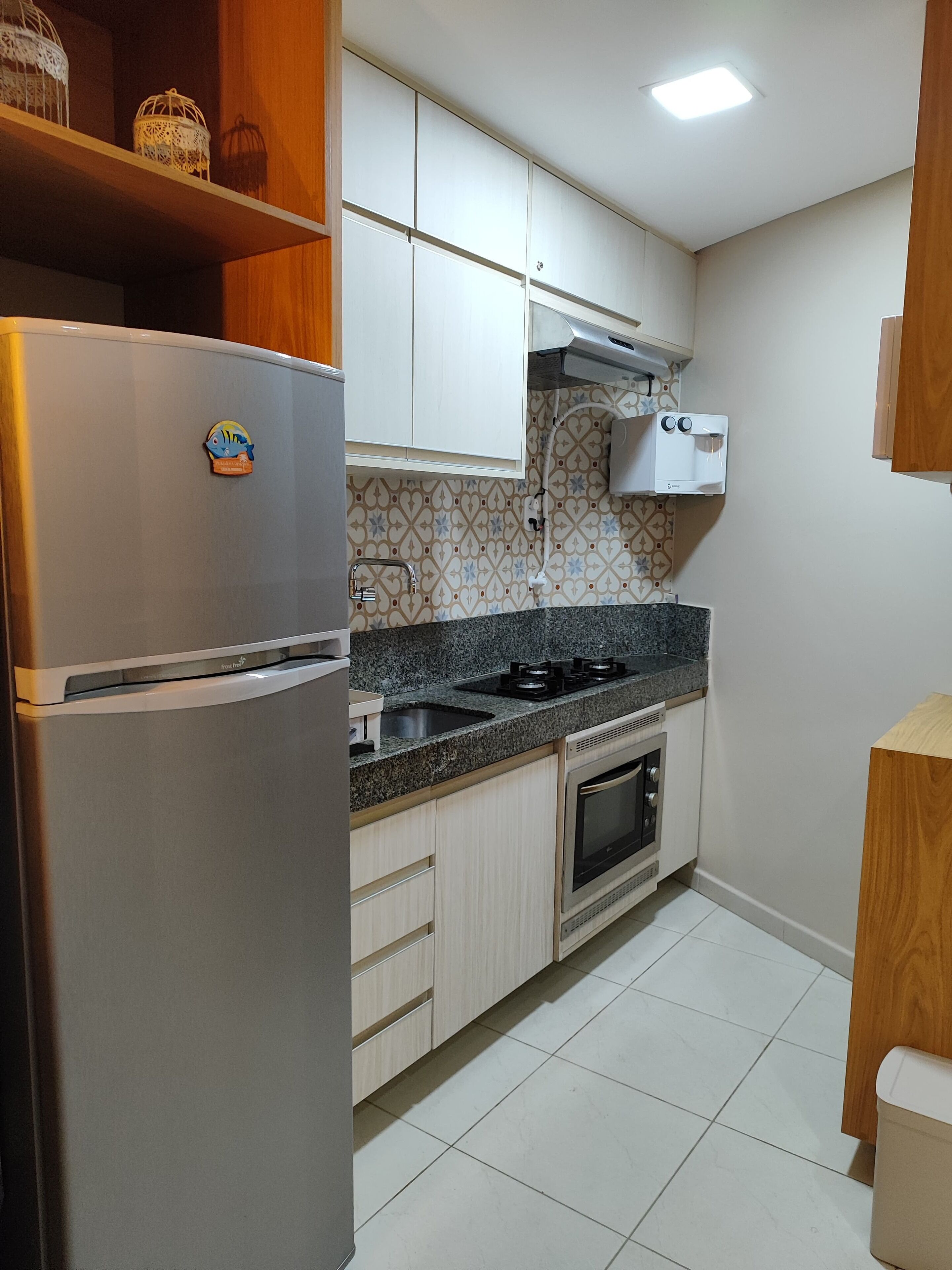 A kitchen with a stainless steel refrigerator, a gas stove, a built-in oven, and a microwave. There is a wooden shelf above the refrigerator and a tiled backsplash.