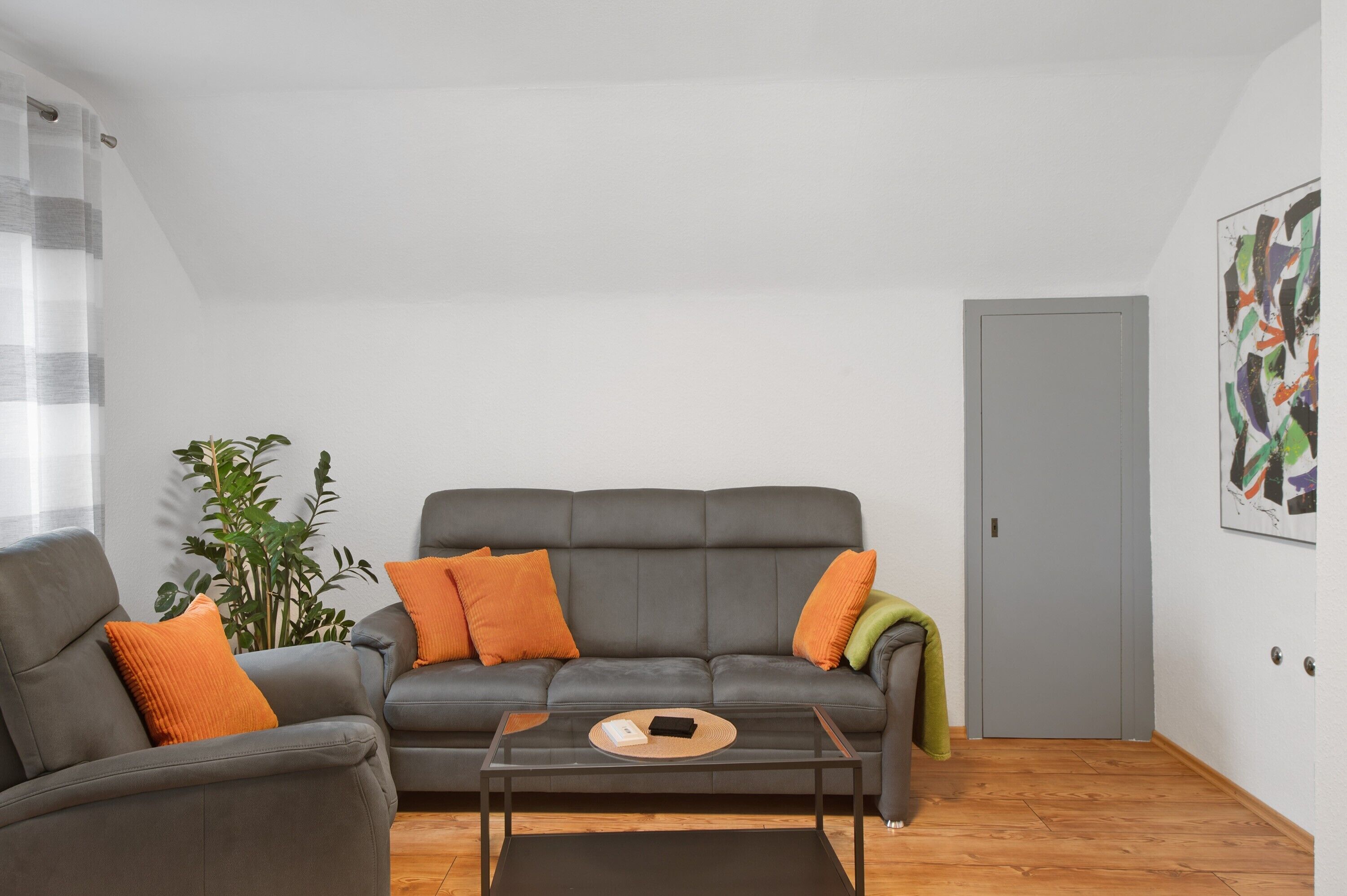 A living room with a grey sofa, two orange cushions, a glass coffee table, a green throw, and a plant.