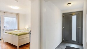 A bedroom with a bed, a window with curtains, a door, and a hallway with a mat. - Apartment 'Kastanie' with Mountain View, Shared Garden and Wi-Fi (Müllheim)