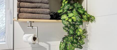 A bathroom with a toilet paper holder, a shelf with towels, and a potted plant.