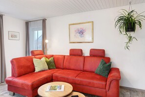 Een rode leren bank met groene kussens, een gekaderd bloemwerk en een hangplant in een kamer met witte wanden en een houten plafond.
