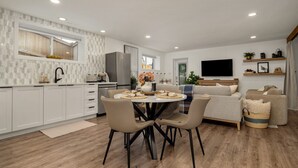 A modern kitchen with white cabinets, a dining table with chairs, and a living area with a sofa and TV. - king/Hot Tub/full kitchen/private Backyard/BBQ/W/D (Lethbridge)