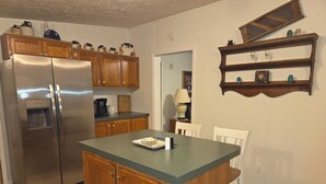 Una cocina con armarios de madera, un refrigerador de acero inoxidable, una pequeña isla con sillas y un estante con objetos decorativos.