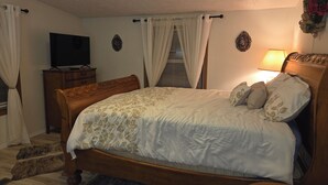 Un dormitorio con una cama de madera, un televisor sobre un mueble bajo, una mesita de noche con lámpara y un adorno en la pared.