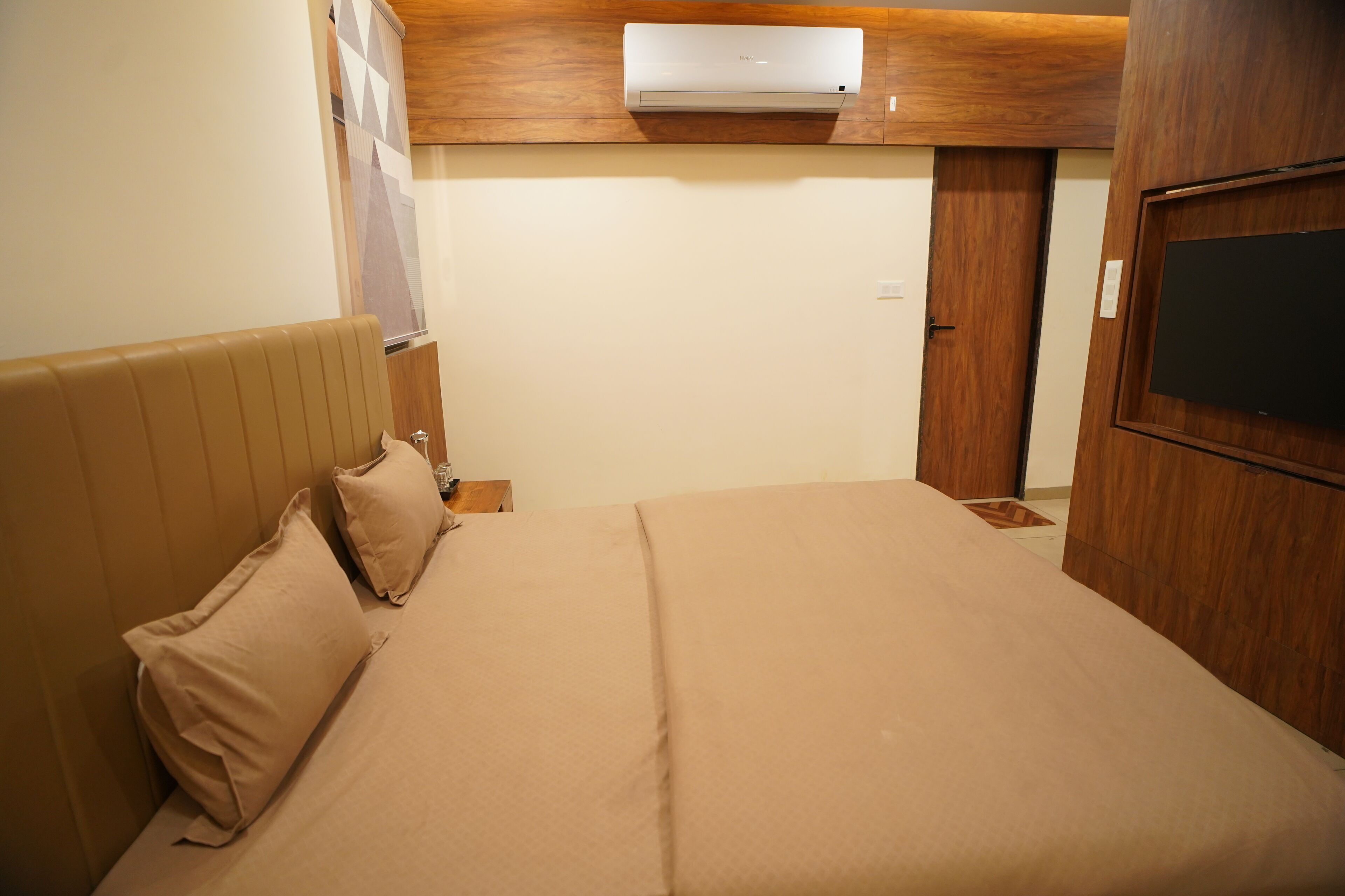 Una habitación de hotel con una cama, almohadas, cabecera de madera, aire acondicionado de pared, una puerta y un televisor en la pared.