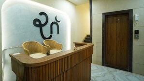 A reception desk with a modern design, two wicker chairs, a wooden counter, and a wooden door with a digital display. - RAAHI HOMESTAY (Ujjain)