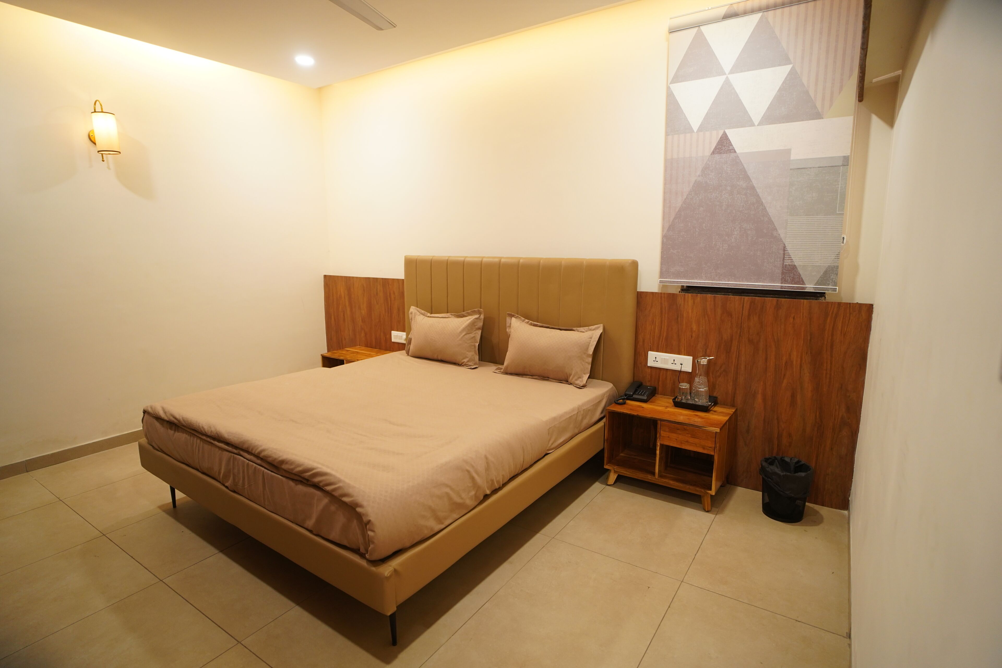A neatly arranged bedroom with a large bed, bedside tables, a wall-mounted light, and a geometric-patterned window.