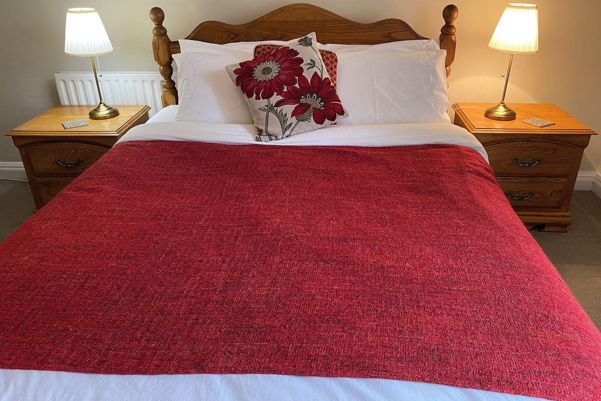 A bed with a red blanket, white pillows, and a floral pillow. There are two bedside tables with lamps, a wooden headboard, and a radiator.