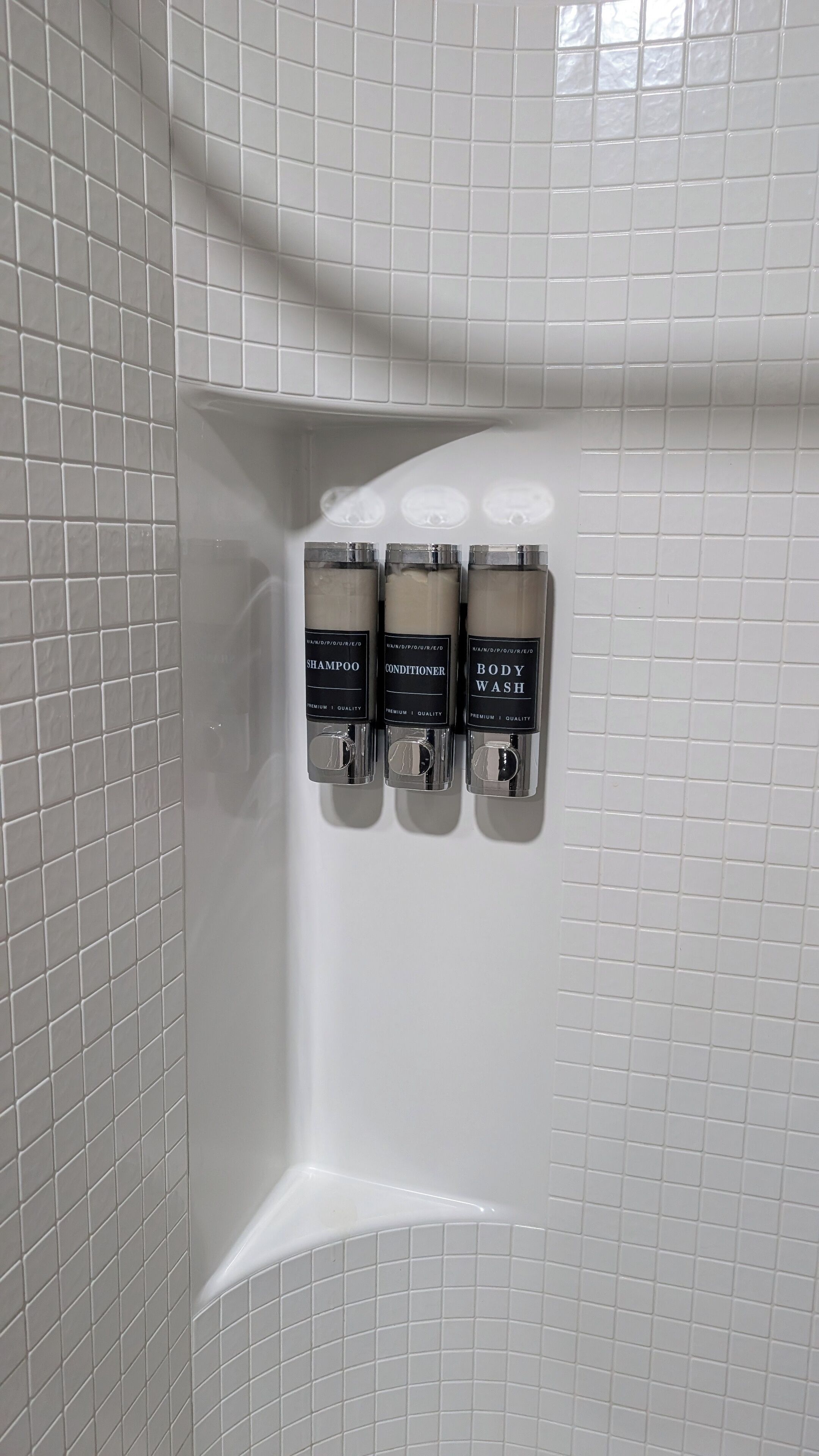 Une salle de bain avec des murs recouverts de carreaux blancs et trois flacons de shampoing, d’après-shampoing et de gel douche fixés au mur.