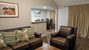 A living room with a leather sofa, a leather armchair, a coffee table, a dining area with a table and chairs, and a kitchen counter with stools.