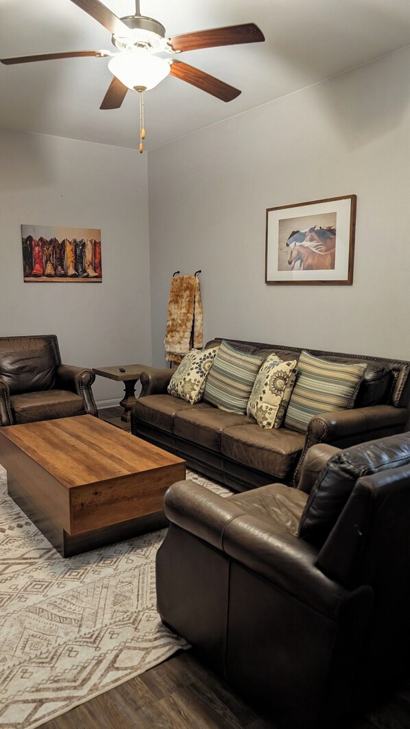 A living room with a brown leather sofa, a wooden coffee table, a ceiling fan, and two framed artworks on the walls. - Secluded Water Front (Sanger)