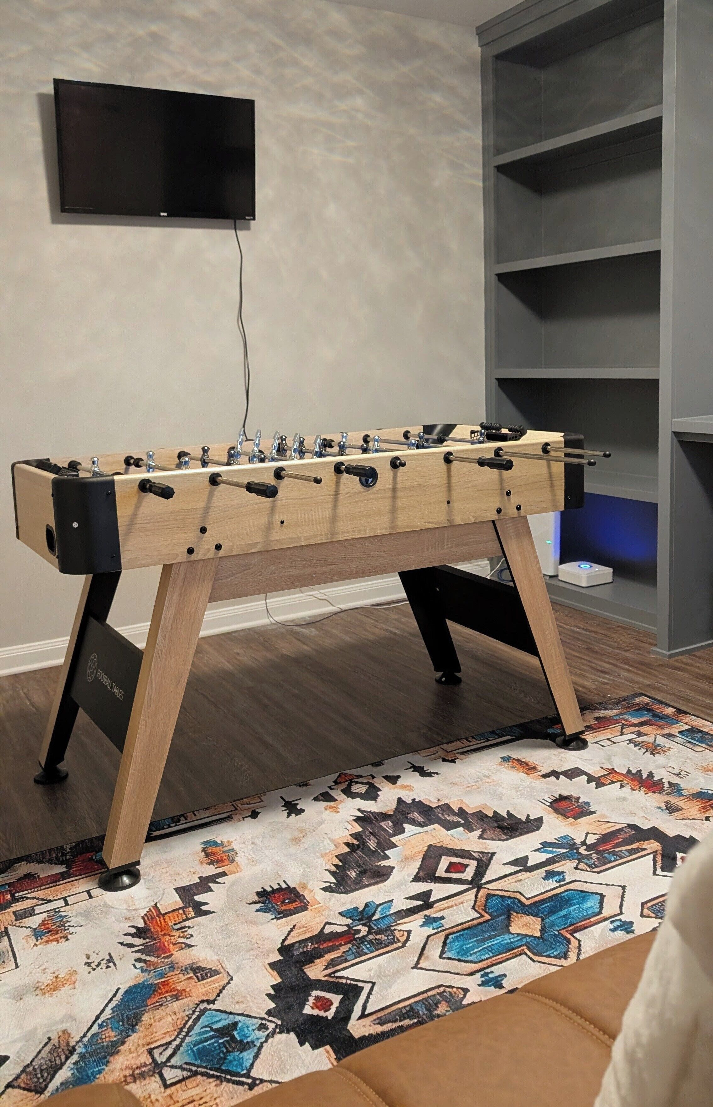 A wooden foosball table with a TV mounted on the wall above it.