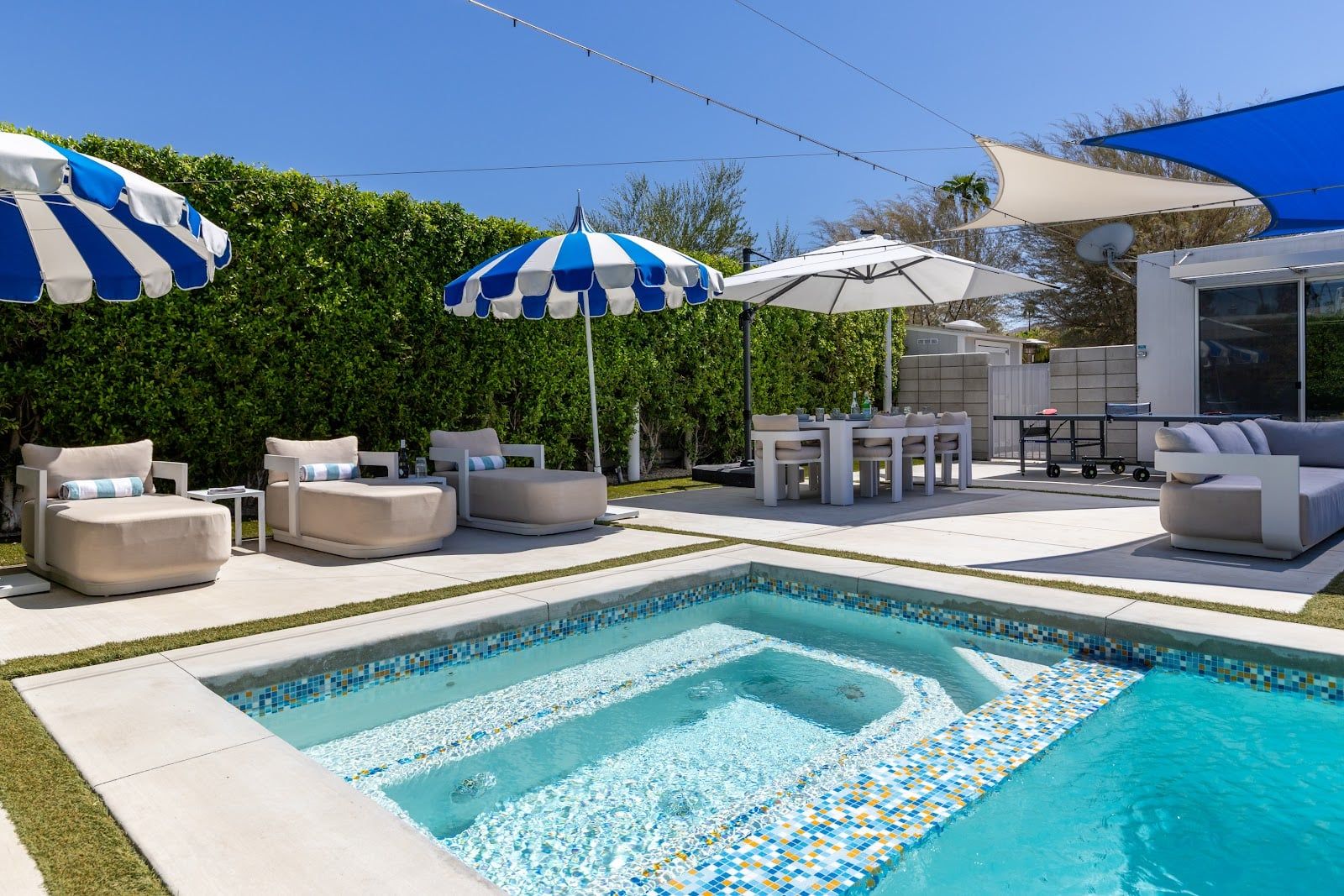 A swimming pool with tiled edges, surrounded by lounge chairs and umbrellas, and a patio area with outdoor furniture.