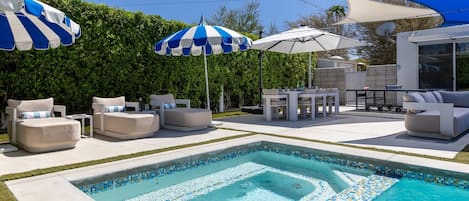 A swimming pool with tiled edges, surrounded by lounge chairs and umbrellas, and a patio area with outdoor furniture.