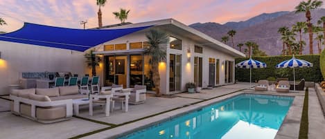 A modern outdoor pool area with a house, lounge chairs, and umbrellas.