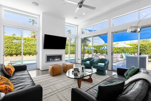 A modern living room with a fireplace, comfortable seating, and large windows offering a view of a pool and greenery. - Modern Desert Oasis – Luxury Retreat (Palm Springs)