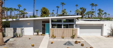A modern two-story house with a garage, surrounded by palm trees and a gravel driveway.